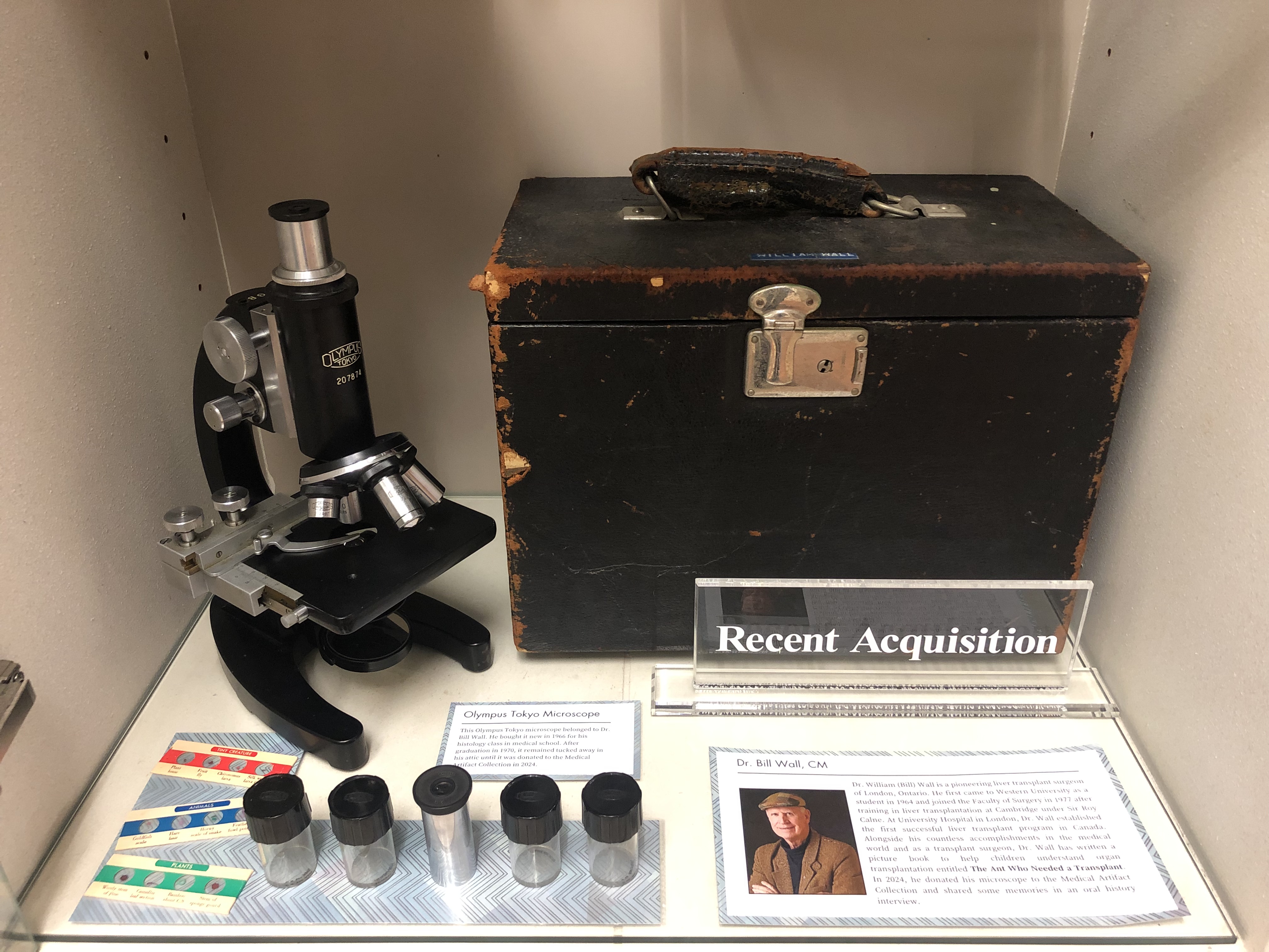 Bottom shelf of display case, featuring Dr. Wall's microscope and case. The microscope is black with four different lenses and one eye piece. The case is hard black leather, and there are extra lenses and containers beside these, as well as slides.